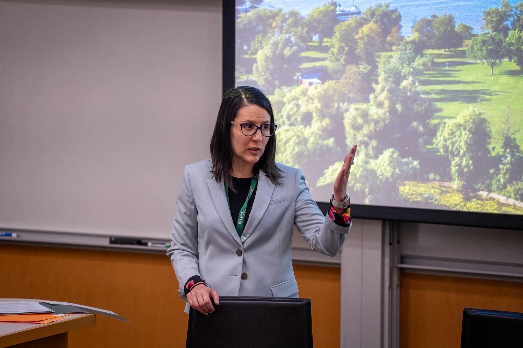 Dr. Zoe Kinias giving the opening address at the Wood Centre Global Symposium.  Zoe stands at the front of a tiered classroom.  Behind her, slightly out of focus is a projection screen.  Zoe is wearing a light grey jacket over a dark pant suit and is holding onto a high stool with her  right hand