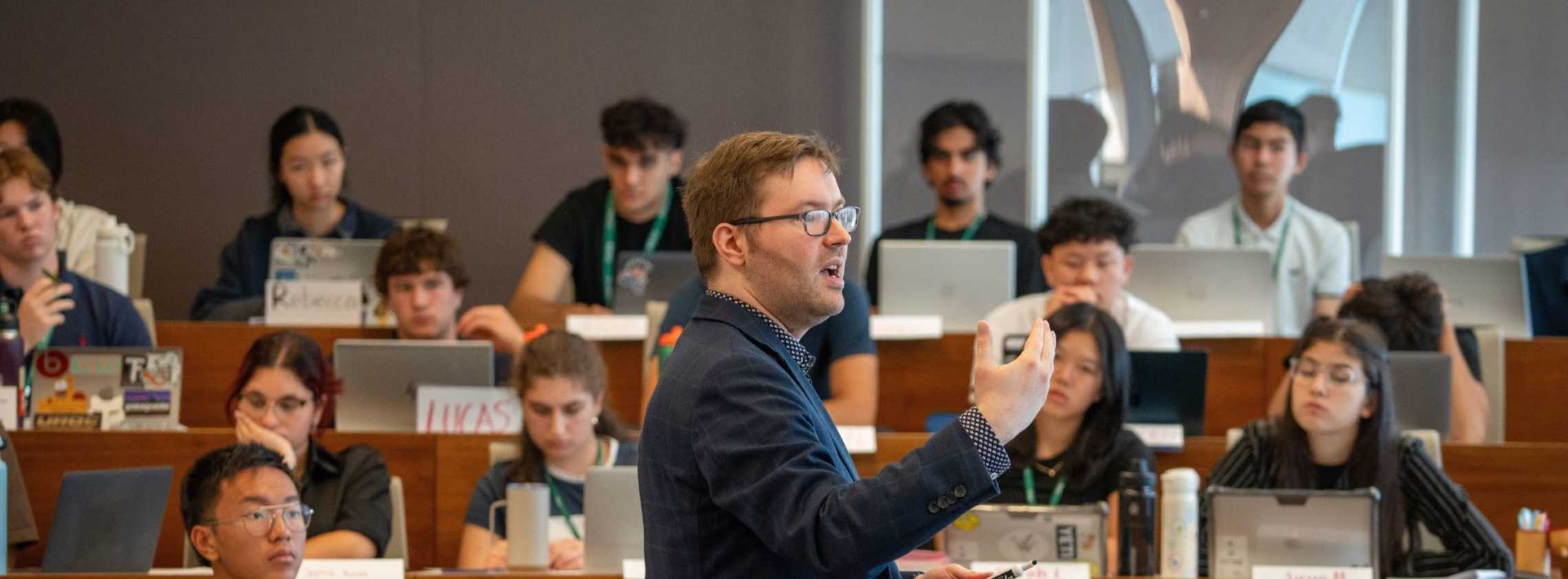 A tiered classroom full of students. The students are paying attention to the Professor in the middle of the room.