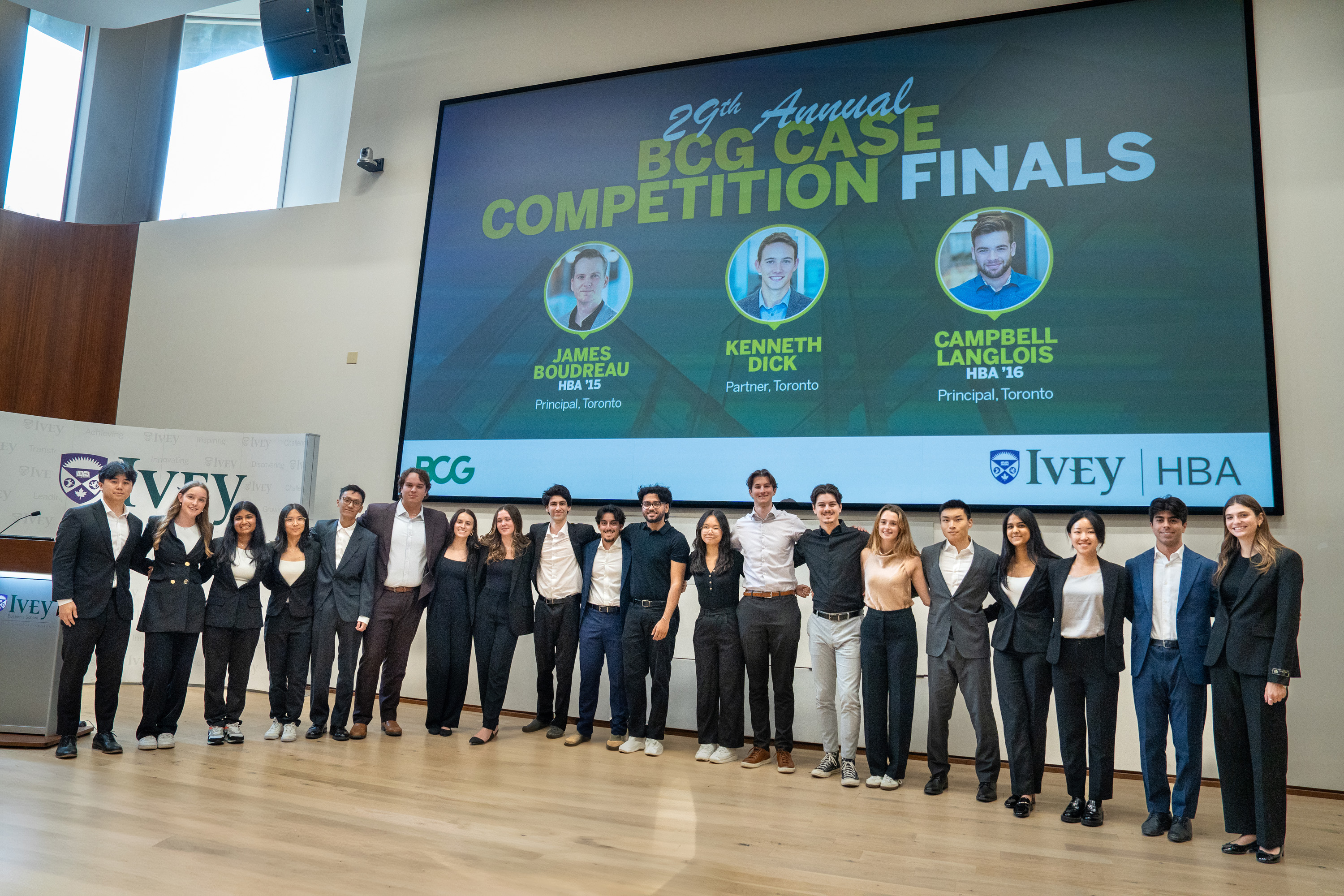 20 students in business attire stand on the stage of the Ivey auditorium for a group photo with the BCG Case Competition slide featuring the finalists judges behind them