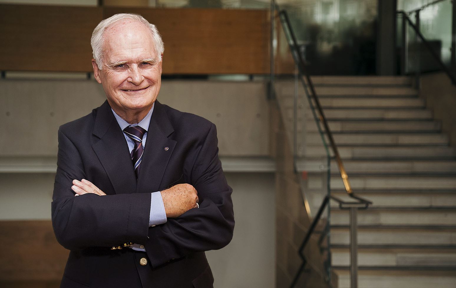 John F. Wood stood with his arms folded in front of the main staircase at Ivey. Mr. Wood is wearing a dark suit and is smiling.