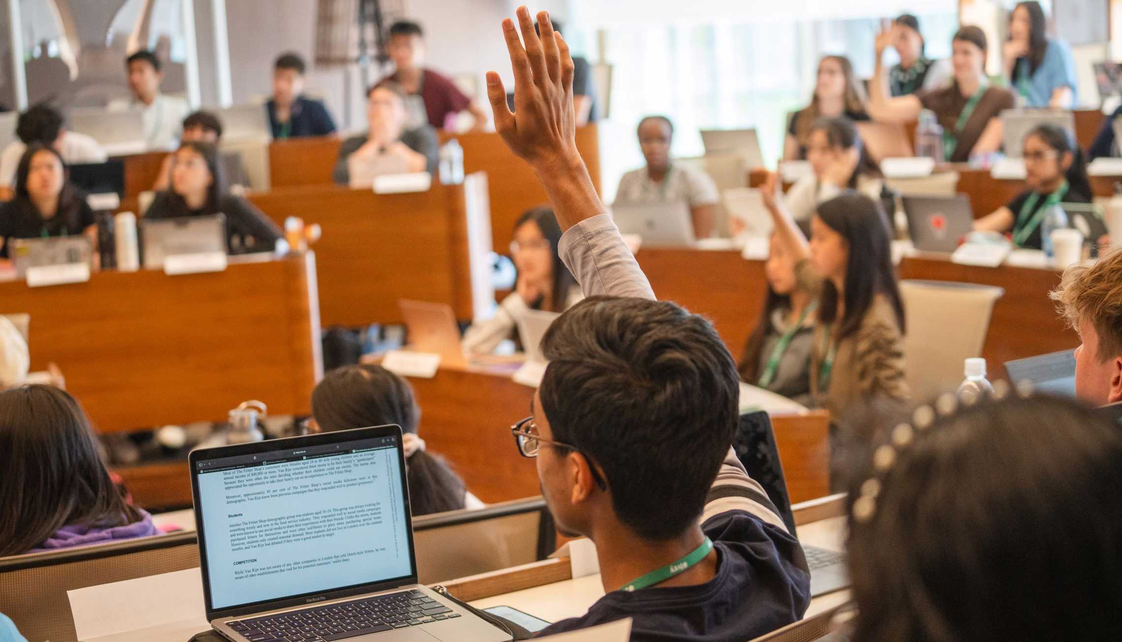 A tiered classroom full of high school students with their hands raised to answer a question.  Most of the students have a laptop on the desk in front of them.