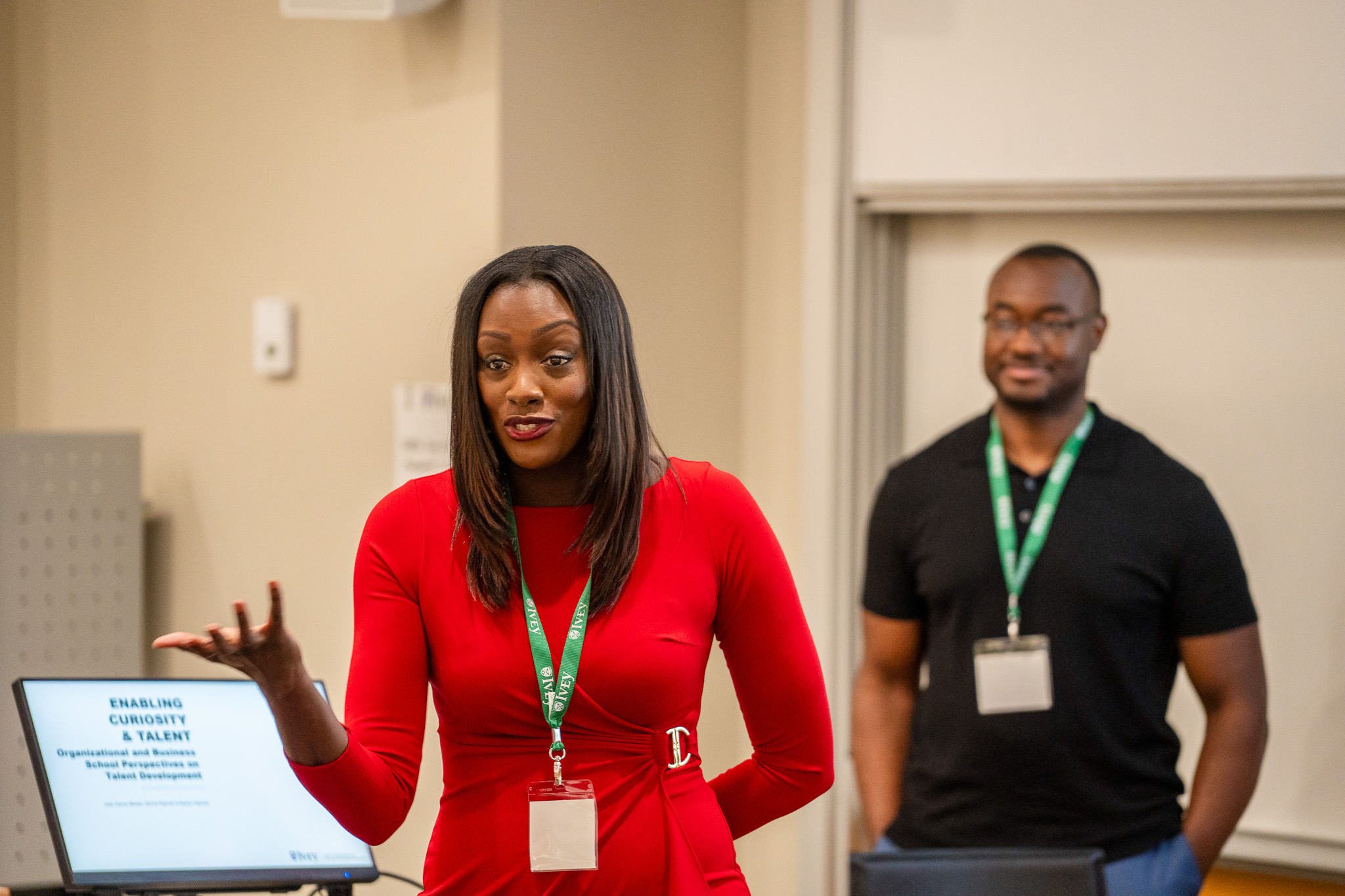 ICON Talent partners cofounders Marilyn & Derrick Raphael presenting at the Wood Symposium 2024.  Marylin is in the front of the photo, wearing a red dress, leading an interactive session.  Derrick is in the background, slightly out of focus, he is wearing jeans and a black teeshirt, has his hands in his pockets and is smiling in agreement with Marilyn's comments.